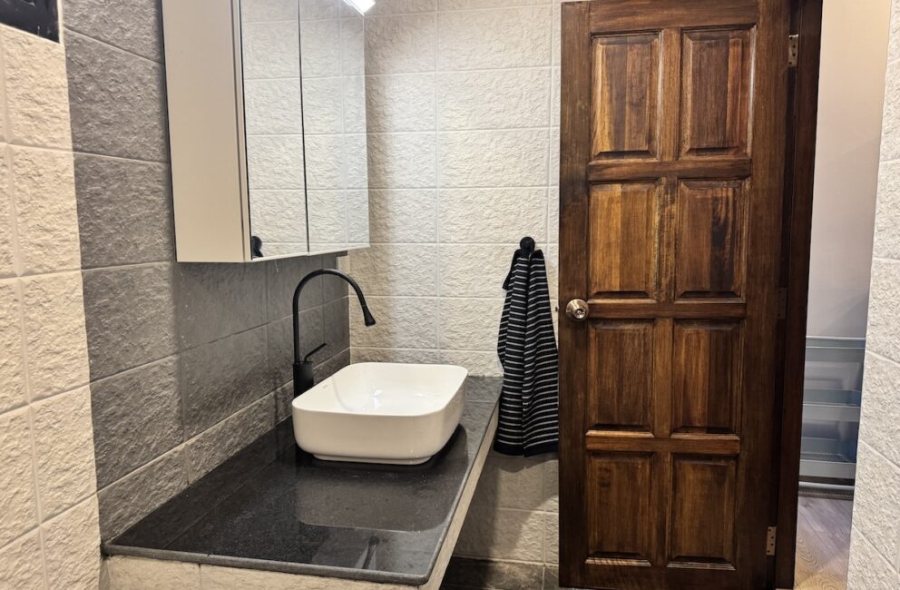 Bathroom with a white vessel basin on a black granite counter, black tap, mirror cabinet and a wooden door in Secret Garden Pool Villa B2